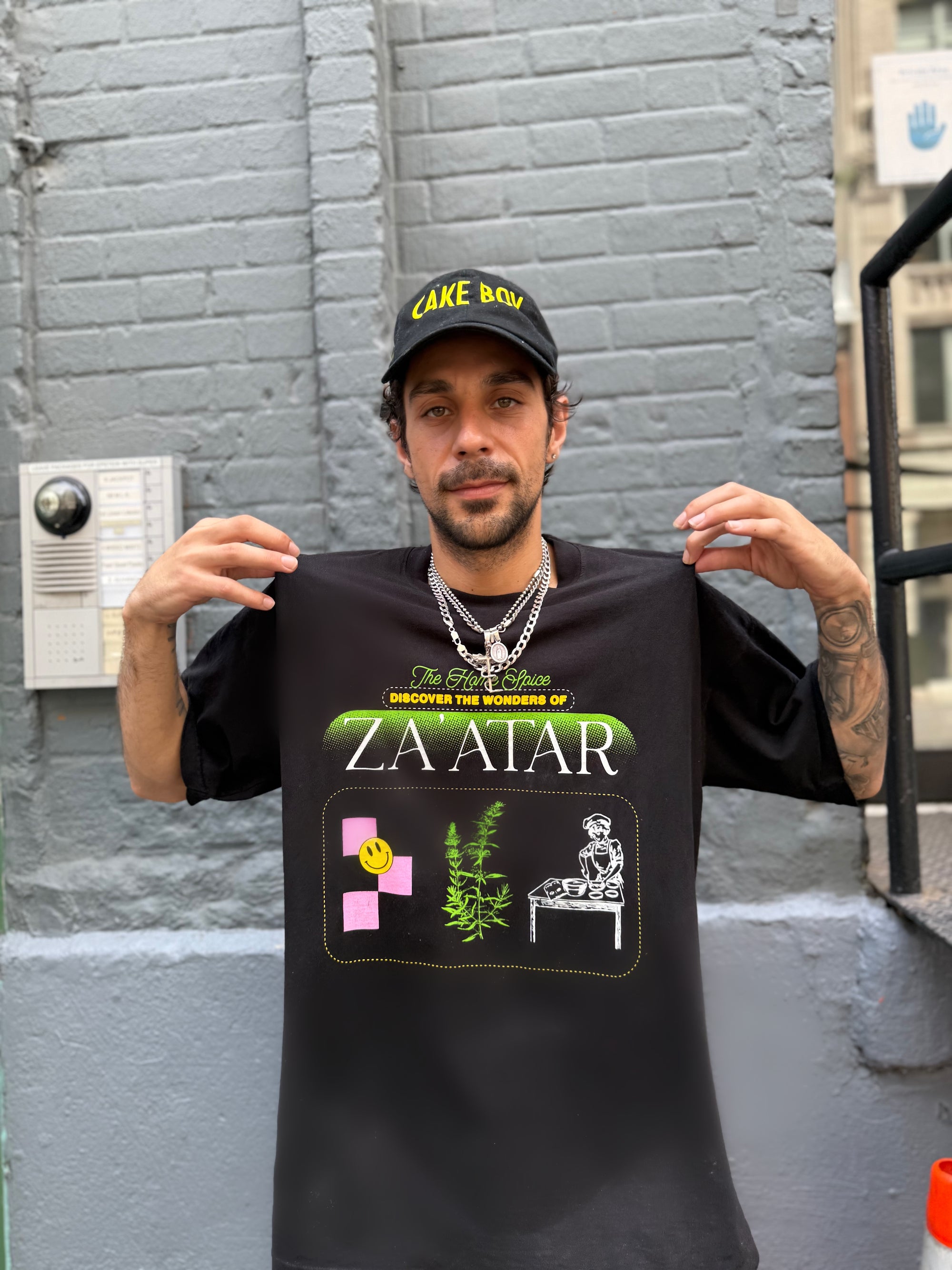 Man holding a black t-shirt with graphic designs and text against a gray wall.